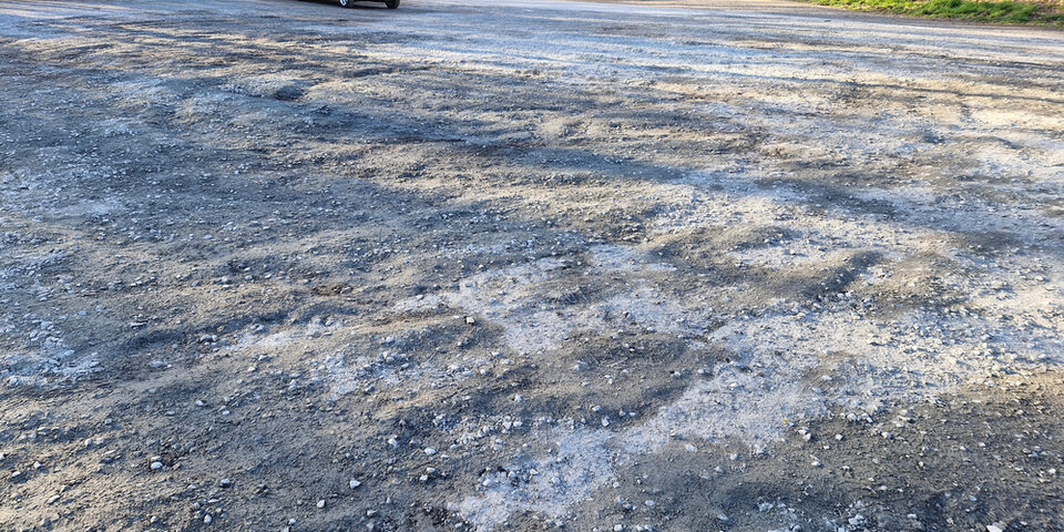 Parkplatz Lauftreff mit beschädigter Schotter-Decke. Im Hintergrund parkt ein weißes Auto.