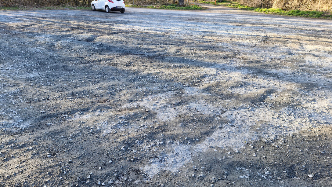 Parkplatz Lauftreff mit beschädigter Schotter-Decke. Im Hintergrund parkt ein weißes Auto.
