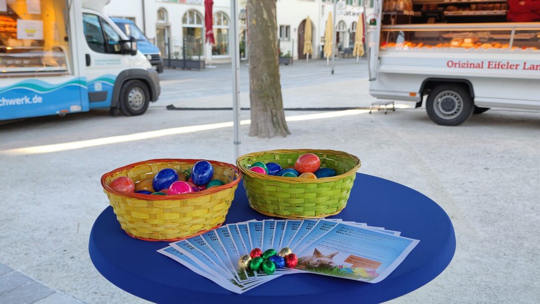Körbchen mit bunten Ostereiern und Flyer zur Osteraktion liegen auf Stehtisch. Im Hintergrund sieht man zwei Verkaufswagen des Wochenmarktes auf dem Marktplatz.