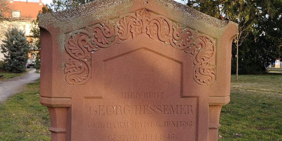 Grabstein mit der Aufschrift "Hier ruht Georg Hessemer". Im Hintergrund grüner Rasen und Bäume.