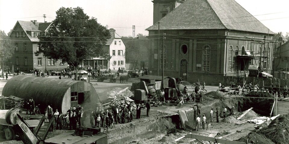 Ein Schwarz-Weiß Bild welches den Marktpaltz in Rüsselsheim am Main zeigt. Im Vordergrund ist ein provisorisches Gebäude und verschiedene Menschen arbeiten an Baugeräten.