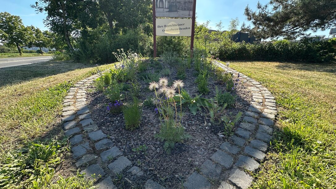 Ortseingang von Bauschheim mit Schild und wappenförmig verlegte Kopfsteine um Blumen.