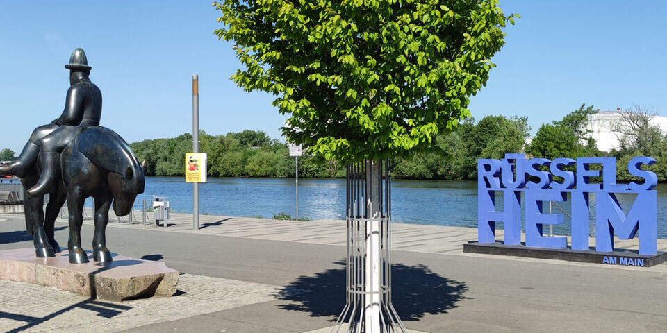 Schriftzug "Rüsselsheim am Main" am Landungsplatz am Main. Links daneben das Leinreiter-Denkmal und ein grün blühender Baum.