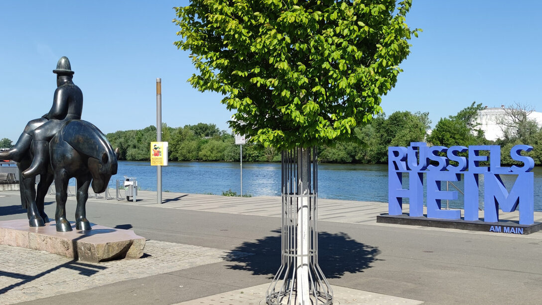 Schriftzug "Rüsselsheim am Main" am Landungsplatz am Main. Links daneben das Leinreiter-Denkmal und ein grün blühender Baum.