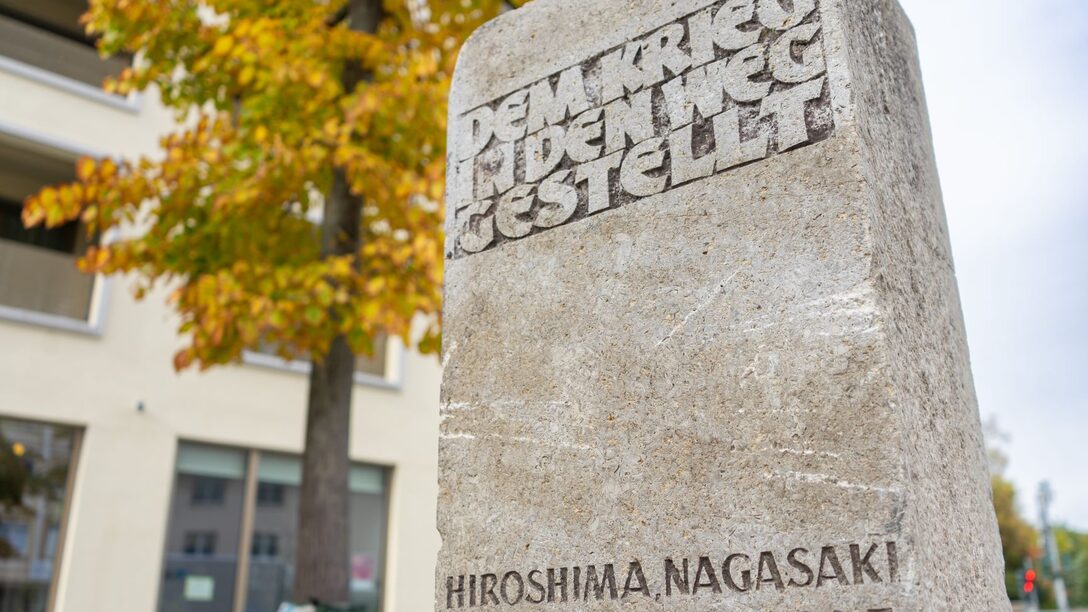 Friedensstein mit der Aufschrift "Dem Krieg in den Weg gestellt. Hiroshima, Nagasaki 6./.9.8.1945" auf dem Friedensplatz. Im Hintergrund ein herbstlich-bunt blühender Baum.