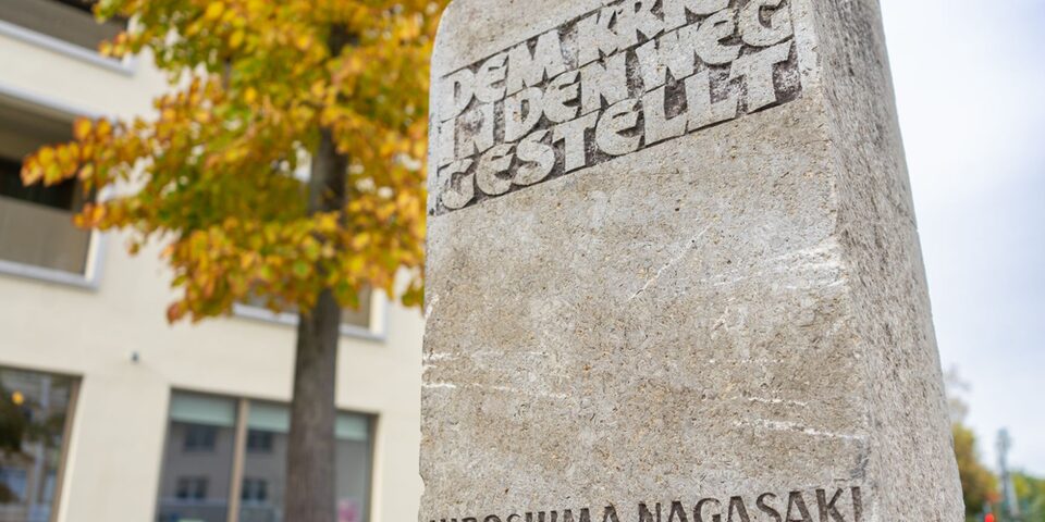 Friedensstein mit der Aufschrift "Dem Krieg in den Weg gestellt. Hiroshima, Nagasaki 6./.9.8.1945" auf dem Friedensplatz. Im Hintergrund ein herbstlich-bunt blühender Baum.
