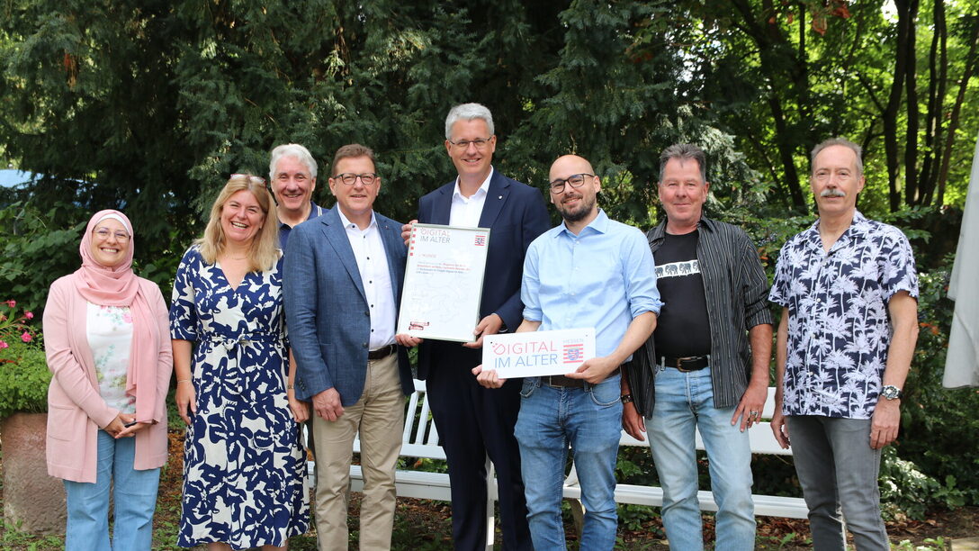 Mehrere Personen stehen vor einer Bank und Bäumen. Zwei halten ein Zertifikat für den Dia-Lotsen-Stützpunkt sowie das dazugehörige Schild in den Händen.