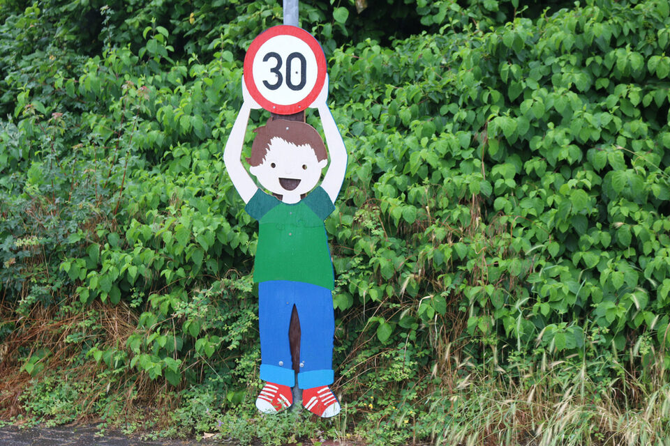 Gemaltes Schild: Kind hält "30er"-Verkehrsschild in die Höhe. Dahinter grüne Büsche.