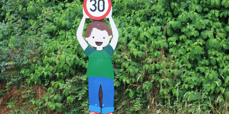 Gemaltes Schild: Kind hält "30er"-Verkehrsschild in die Höhe. Dahinter grüne Büsche.
