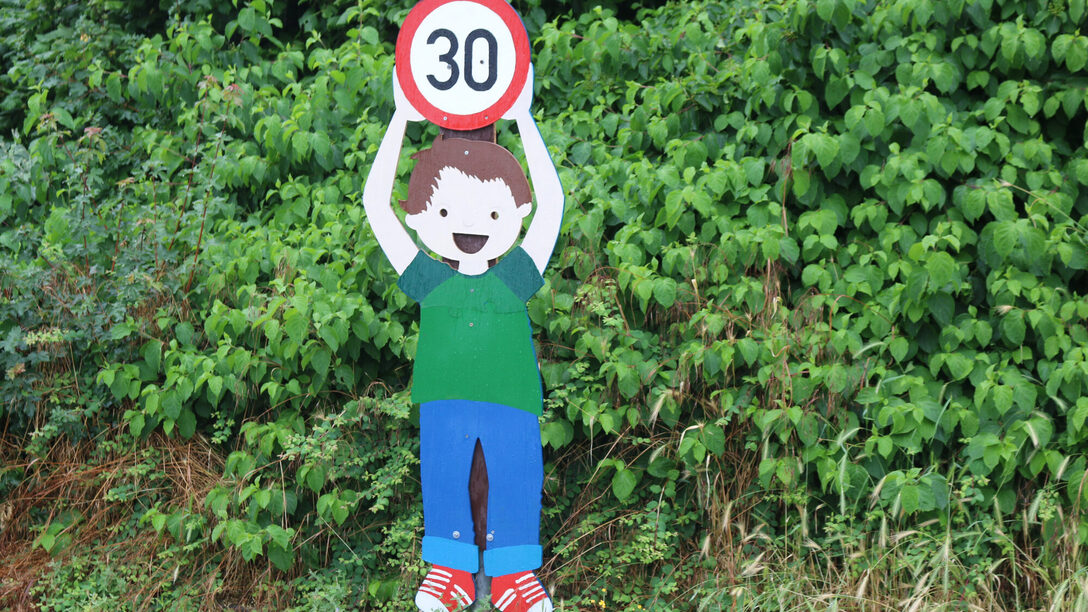 Gemaltes Schild: Kind hält "30er"-Verkehrsschild in die Höhe. Dahinter grüne Büsche.