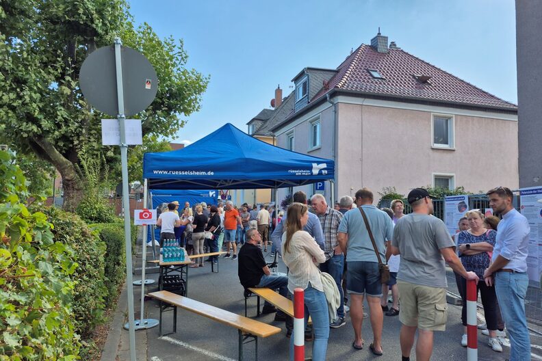 Menschen stehen auf der Straße. Im Hintergrund ein blauer "Rüsselsheim"-Pavillion, Bierzeltgarnituren und Getränke.
