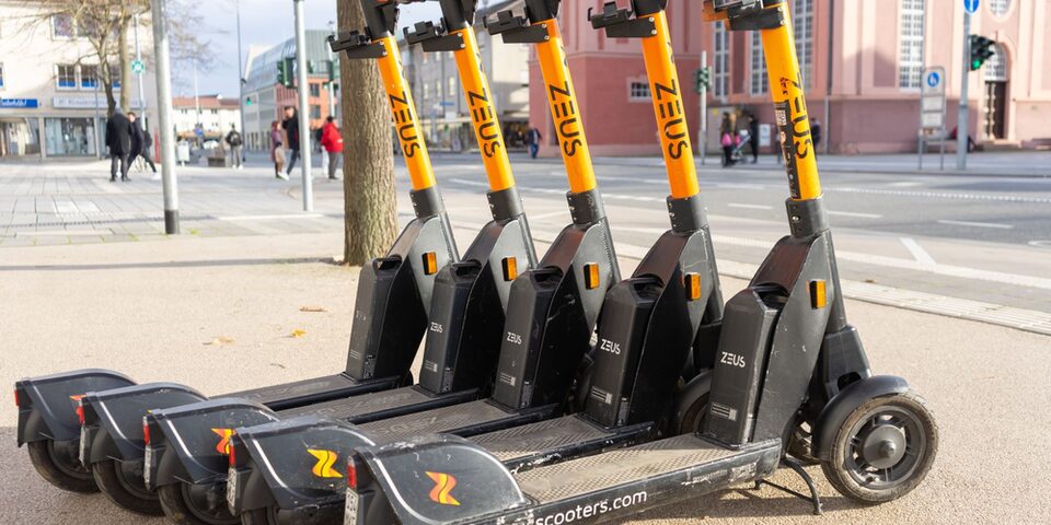Mehrere E-Scooter der Firma "ZEUS" in orange-schwarz stehen an der Ecke Mainstraße/Frankfurter Straße. Im Hintergrund die Stadtkirche.
