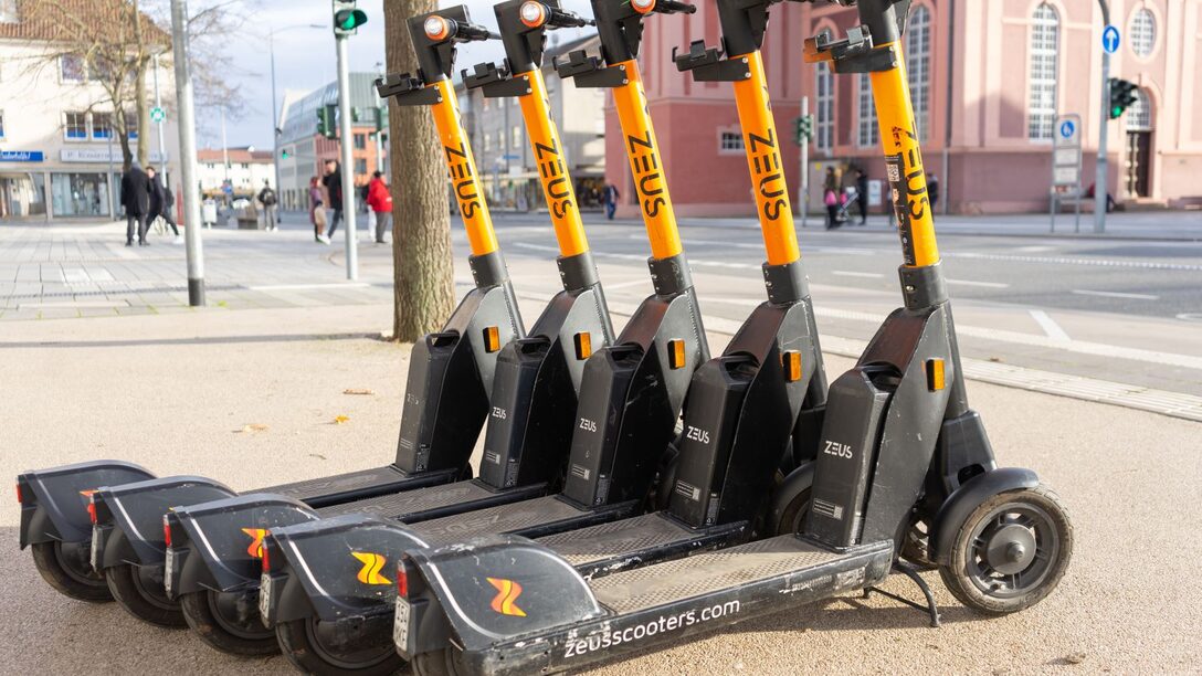 Mehrere E-Scooter der Firma "ZEUS" in orange-schwarz stehen an der Ecke Mainstraße/Frankfurter Straße. Im Hintergrund die Stadtkirche.