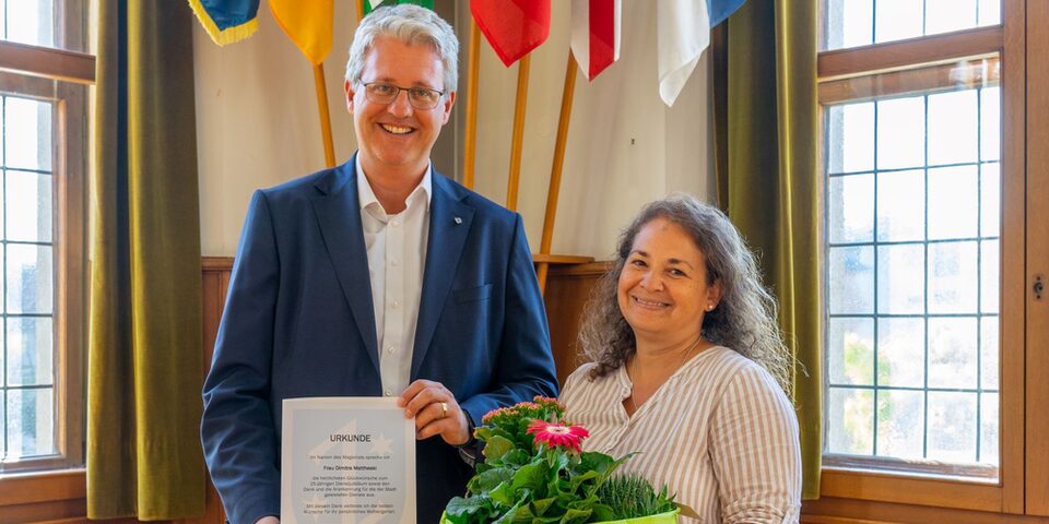 Oberbürgermeister Patrick Burghardt hält eine Urkunde, neben ihm Dimitra Mattheaki mit einem Blumenstrauß in den Händen