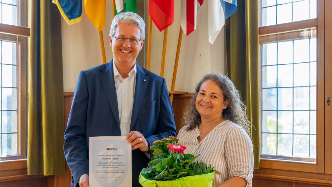 Oberbürgermeister Patrick Burghardt hält eine Urkunde, neben ihm Dimitra Mattheaki mit einem Blumenstrauß in den Händen