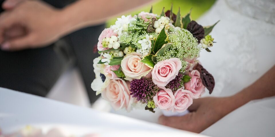 Hände halten einen Blumenstrauß, im Hintergrund ist der Ausschnitt eines sitzenden Brautpaares angedeutet.