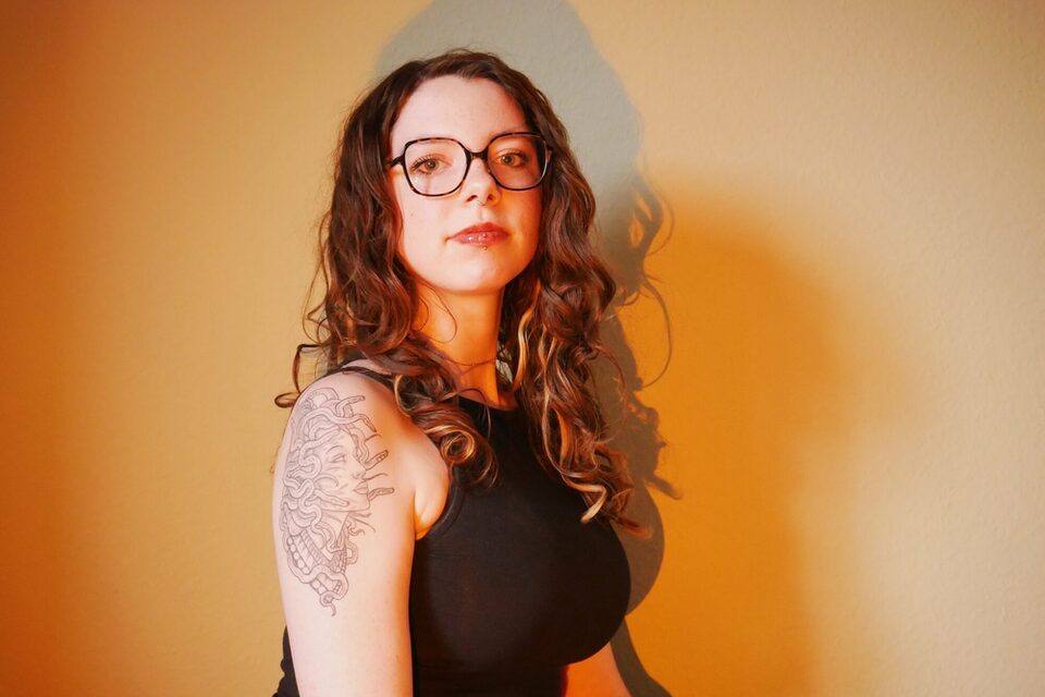 Eine Person sitzt vor einer orange ausgeleuchteten Wand und blickt in die Kamera. Sie trägt eine Brille. Auf dem Arm ist ein Tatoo eines Medusa-Kopfes.
