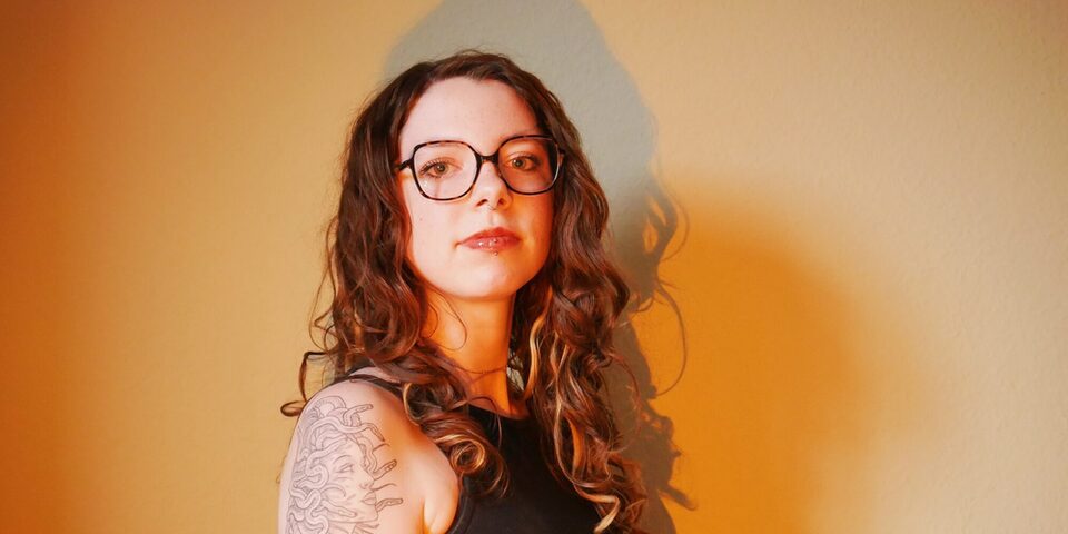 Eine Person sitzt vor einer orange ausgeleuchteten Wand und blickt in die Kamera. Sie trägt eine Brille. Auf dem Arm ist ein Tatoo eines Medusa-Kopfes.