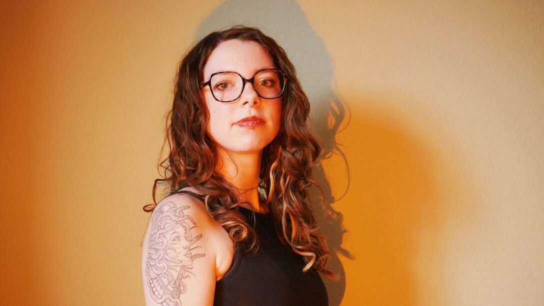 Eine Person sitzt vor einer orange ausgeleuchteten Wand und blickt in die Kamera. Sie trägt eine Brille. Auf dem Arm ist ein Tatoo eines Medusa-Kopfes.