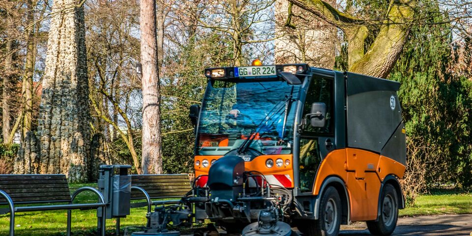 Kehrmaschine des Städteservice kehrt einen Weg auf einer Grünfläche. Im Hintergrund Bäume und zwei Sitzbänke bei sonnigem Wetter.