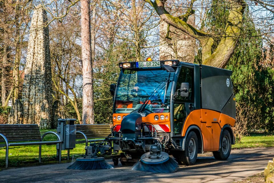 Kehrmaschine des Städteservice kehrt einen Weg auf einer Grünfläche. Im Hintergrund Bäume und zwei Sitzbänke bei sonnigem Wetter.