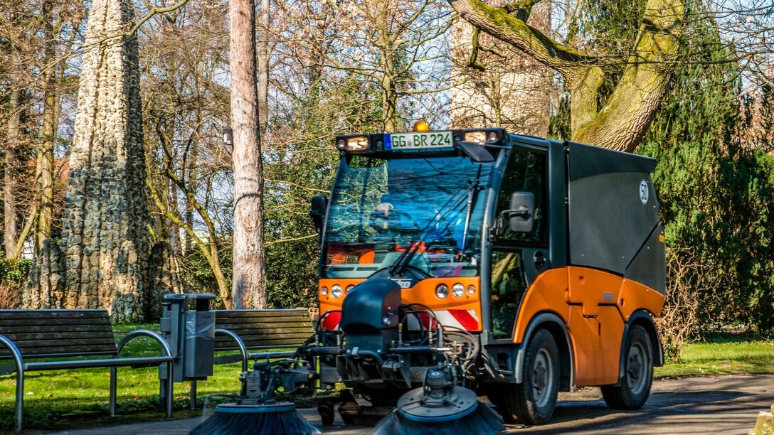 Kehrmaschine des Städteservice kehrt einen Weg auf einer Grünfläche. Im Hintergrund Bäume und zwei Sitzbänke bei sonnigem Wetter.