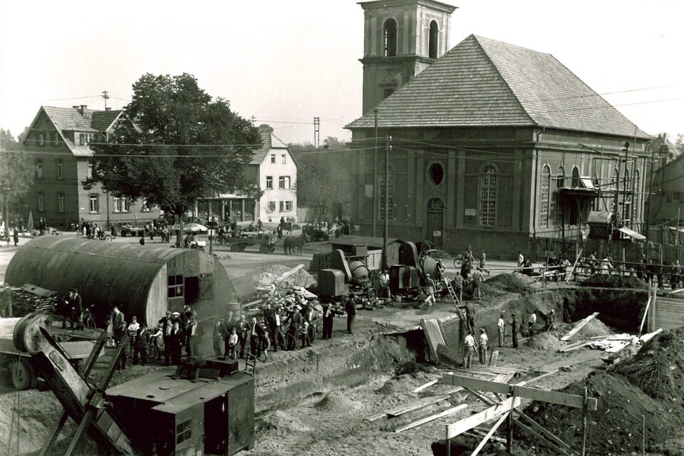 Schwarz-Weiß-Foto des Marktplatz in Rüsselsheim. Im Hintergrund die Stadtkirche, im Vordergrund stehen mehrere Menschen auf einer Baustelle mit Zementmischern und einer Barracke auf der linken Seite.
