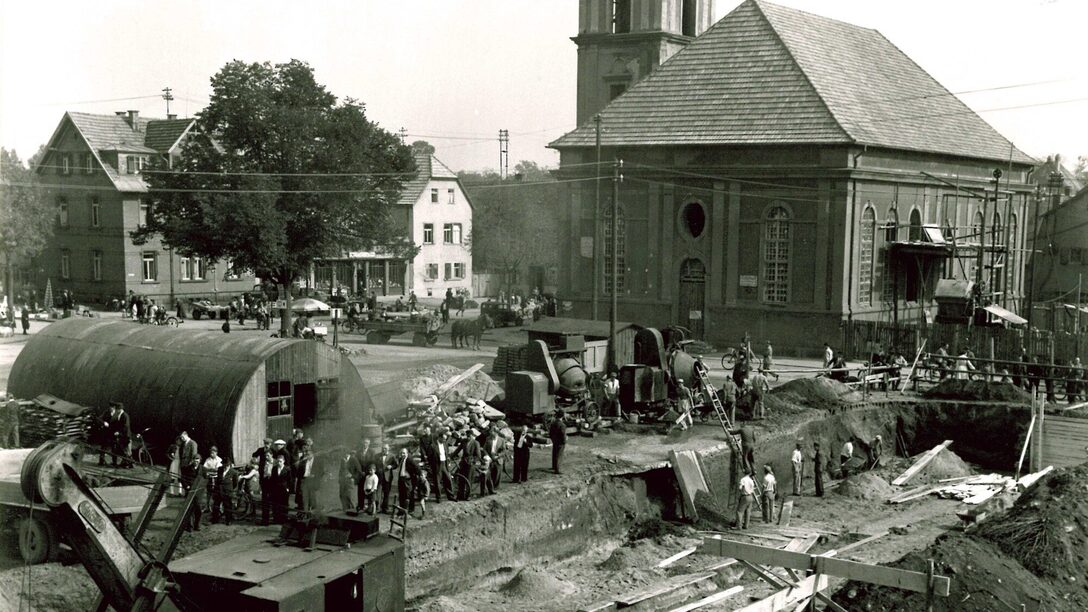 Schwarz-Weiß-Foto des Marktplatz in Rüsselsheim. Im Hintergrund die Stadtkirche, im Vordergrund stehen mehrere Menschen auf einer Baustelle mit Zementmischern und einer Barracke auf der linken Seite.