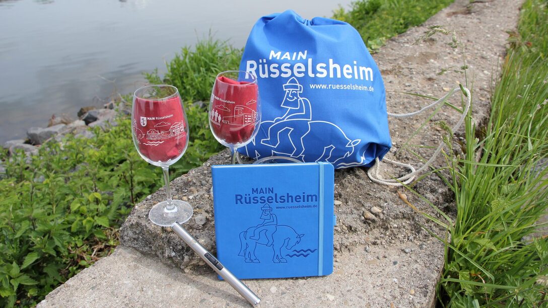 Zwei Weingläser, ein Notizbuch und ein Turnbeutel sind vor dem Main drapiert. Alle Produkte haben die Aufschrift "MAIN Rüsselsheim".
