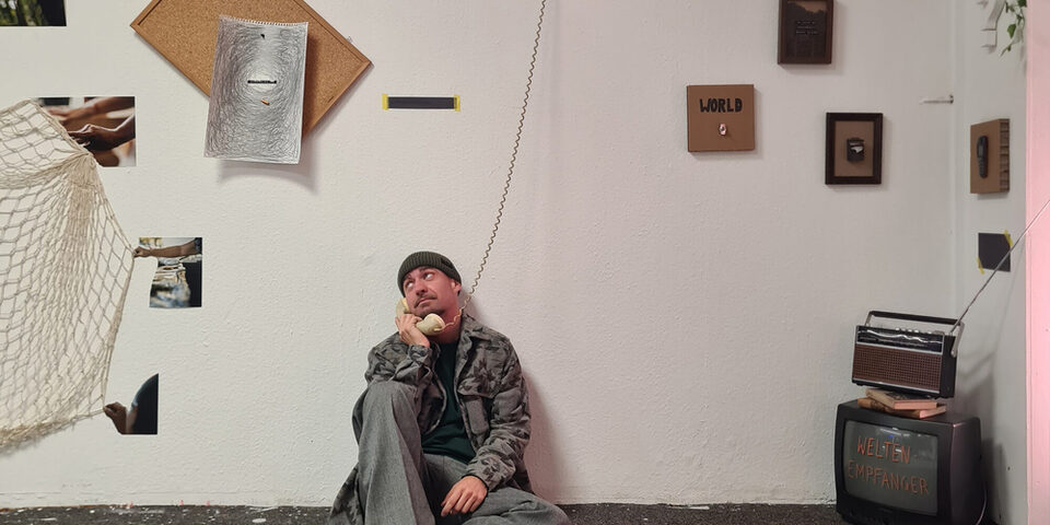 Eine Person sitzt vor einer Wand, an der mehrere Bilder hängen, und hat einen Telefonhörer am Ohr.