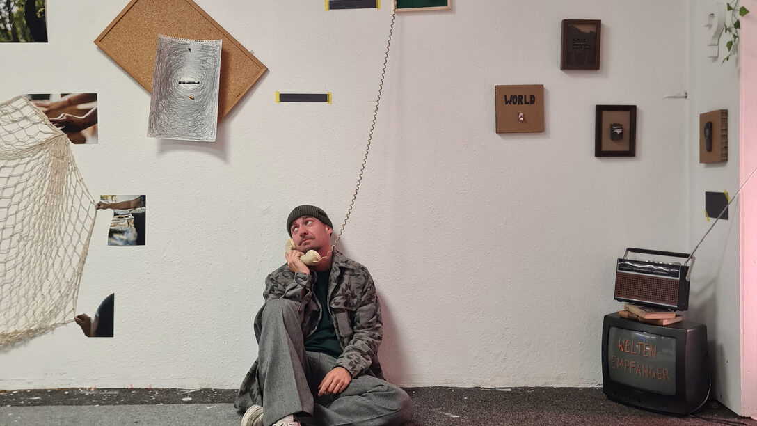 Eine Person sitzt vor einer Wand, an der mehrere Bilder hängen, und hat einen Telefonhörer am Ohr.