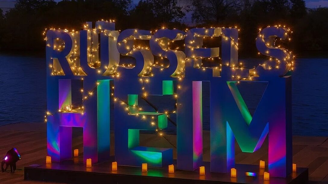 Rüsselsheim-Schriftzug, der mit Lichterketten beleuchtet ist und bunt schimmert. Im Hintergrund im Abenddunkel der Main.