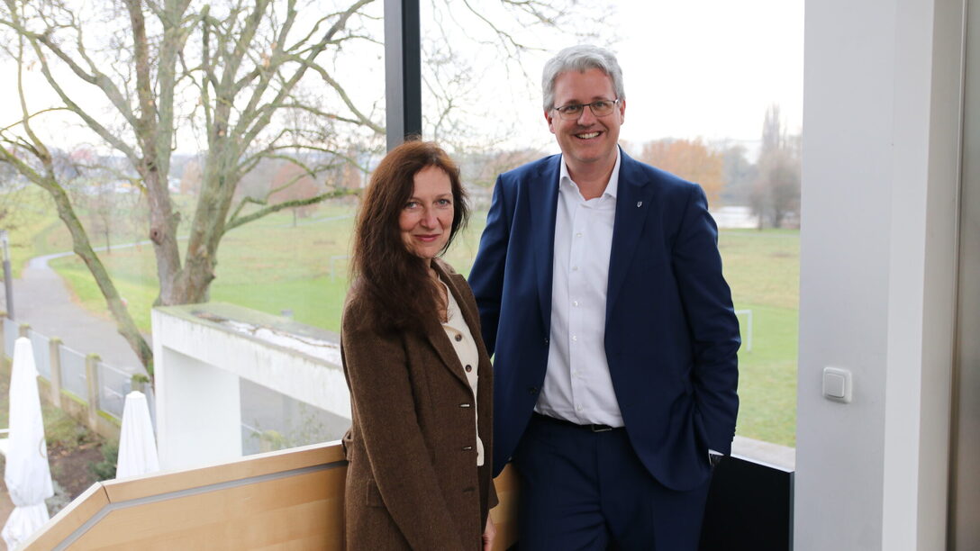 Dr. Beate Kemfert (Vorstand der Stiftung Opelvillen) und Oberbürgermeister Patrick Burghardt stehen vor einem Fenster in den Opelvillen mit Blick auf das grüne Mainvorland.