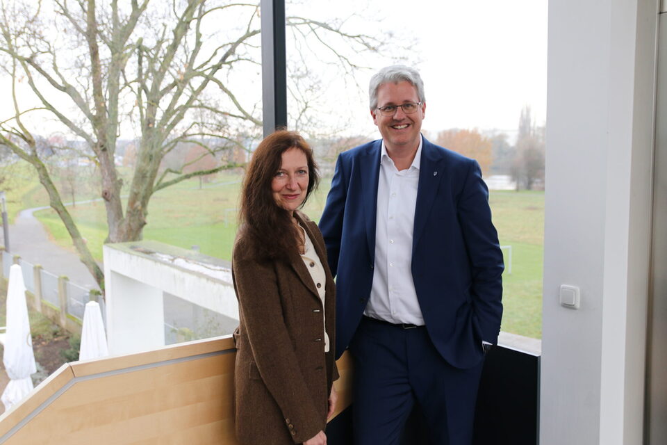 Dr. Beate Kemfert (Vorstand der Stiftung Opelvillen) und Oberbürgermeister Patrick Burghardt stehen vor einem Fenster in den Opelvillen mit Blick auf das grüne Mainvorland.