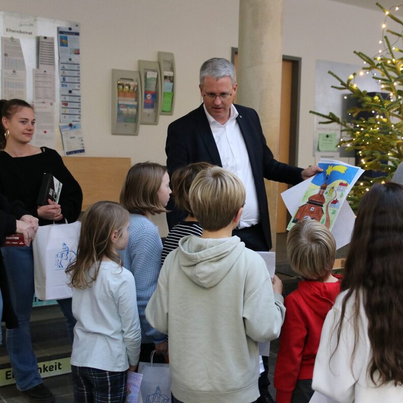 Oberbürgermeister Patrick Burghardt hält ein gemaltes Bild mit Weihnachtsmotiv in der Hand. Vor ihm stehen mehrere Kinder der Ganztagsbetreuung der Eichgrundschule, die man von hinten sieht.