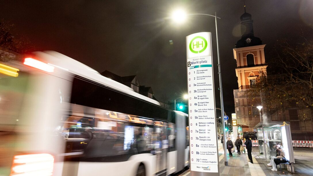 Bushaltestelle "Marktplatz" im Dunkeln. Ein vorbeifahrender Bus, wartende Menschen. Im Hintergrund die Stadtkirche.