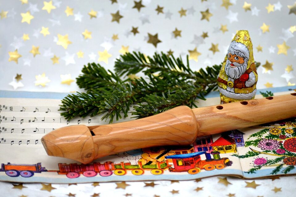 Liederheft mit Noten liegt auf Tischdecke mit Sternen. Darüber ein Tannenzweig, eine Blockflöte und ein Schokoladen-Nikolaus.