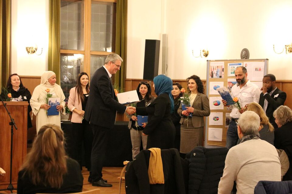 Oberbürgermeister Patrick Burghardt übergibt einer Dame die Urkunde für die Integrationslotsen-Qualifizierung im Trauzimmer. Im Hintergrund stehen bereits andere Integrationslotsinnen und -lotsin mit ihren Urkunden und einer orangenen Rose in der Hand.