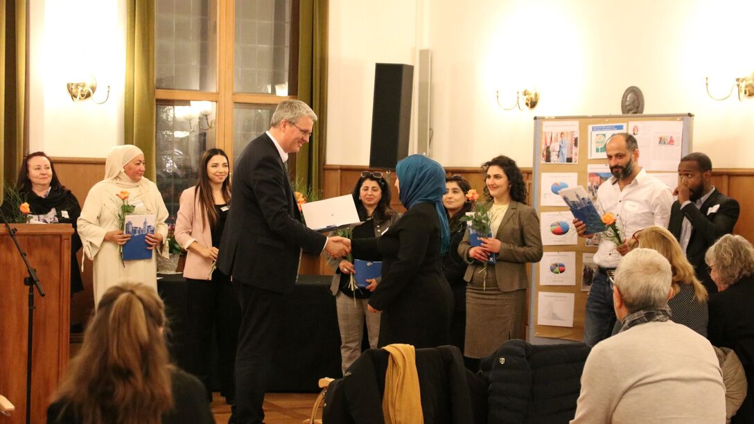 Oberbürgermeister Patrick Burghardt übergibt einer Dame die Urkunde für die Integrationslotsen-Qualifizierung im Trauzimmer. Im Hintergrund stehen bereits andere Integrationslotsinnen und -lotsin mit ihren Urkunden und einer orangenen Rose in der Hand.