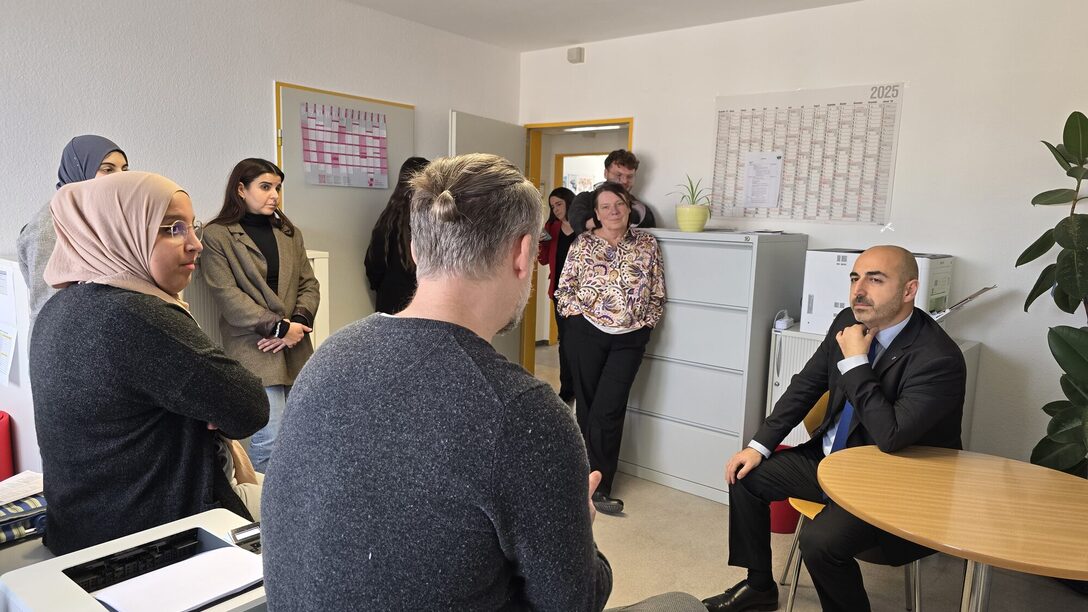 Neun Personen sitzen und stehen in einem Büro und sind in ein Gespräch verwickelt.