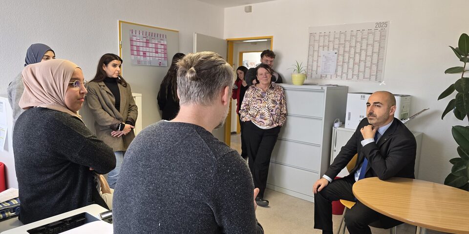 Neun Personen sitzen und stehen in einem Büro und sind in ein Gespräch verwickelt.