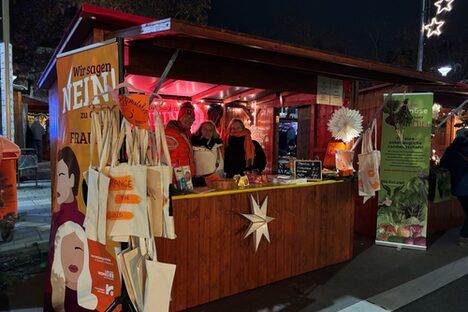 Verkaufshütte auf dem Rüsselsheimer Weihnachtsmarkt, in der drei Personen stehen. An der Seite hängt ein Banner mit der Aufschrift "Wir sagen NEIN zu Gewalt an Frauen"