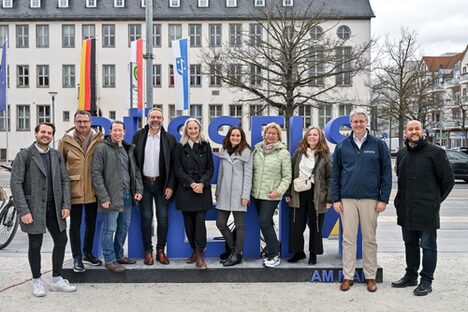 Delegation von Mitarbeitenden der Stabsstelle Kultur und Vielfalt, Oberbürgermeister Patrick Burghardt und weitere stehen vor dem Rüsselsheim Schriftzug auf dem Marktplatz. Im Hintergrund das Rathaus.