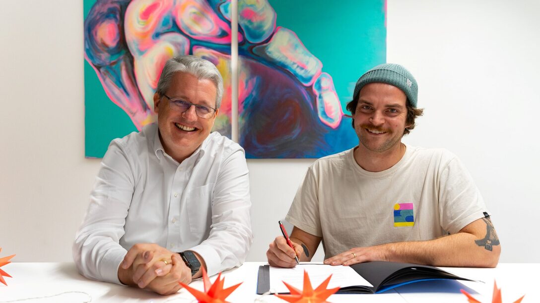 Oberbürgermeister Patrick Burghardt und Benny Becker (Rollwerk) sitzen an einem Tisch und lächeln in die Kamera. Benny Becker hält einen Stift, um die Fördervereinbarung mit der Stadt zu unterzeichnen, die vor ihm auf dem Tisch liegt.