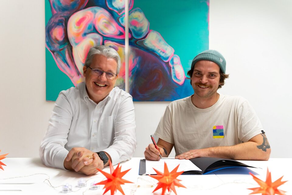 Oberbürgermeister Patrick Burghardt und Benny Becker (Rollwerk) sitzen an einem Tisch und lächeln in die Kamera. Benny Becker hält einen Stift, um die Fördervereinbarung mit der Stadt zu unterzeichnen, die vor ihm auf dem Tisch liegt.