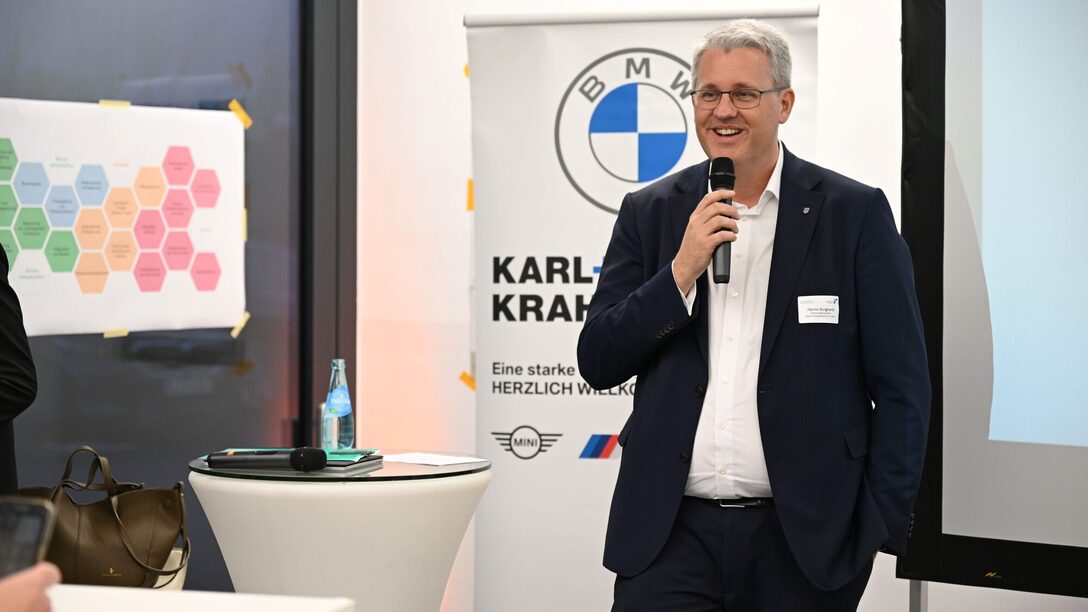 Oberbürgermeister Patrick Burghardt spricht lächelnd in ein Mikrofon. Im Hintergrund ein Rollup des Autohauses Karl+Co. sowie ein Stehtisch und mehrere Poster.