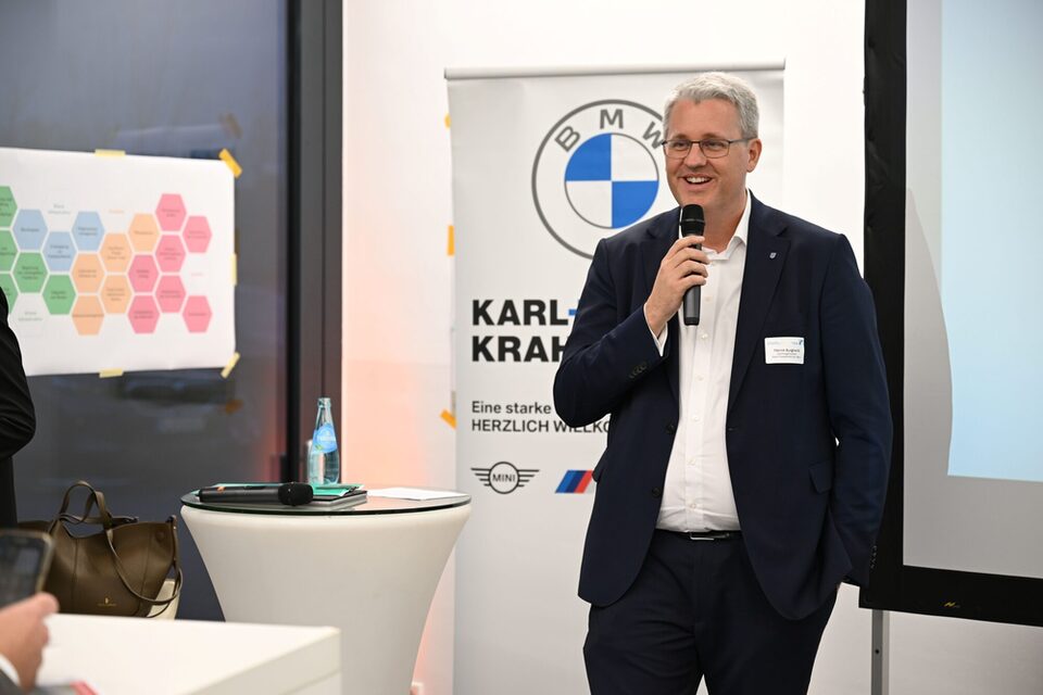 Oberbürgermeister Patrick Burghardt spricht lächelnd in ein Mikrofon. Im Hintergrund ein Rollup des Autohauses Karl+Co. sowie ein Stehtisch und mehrere Poster.