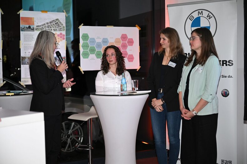 Links im Bild Claudia Gotz, Leiterin der Wirtschaftsförderung, die in ein Mikrofon spricht. An einem Stehtisch stehen drei ehemalige Masterstudentinnen und hören ihr zu.