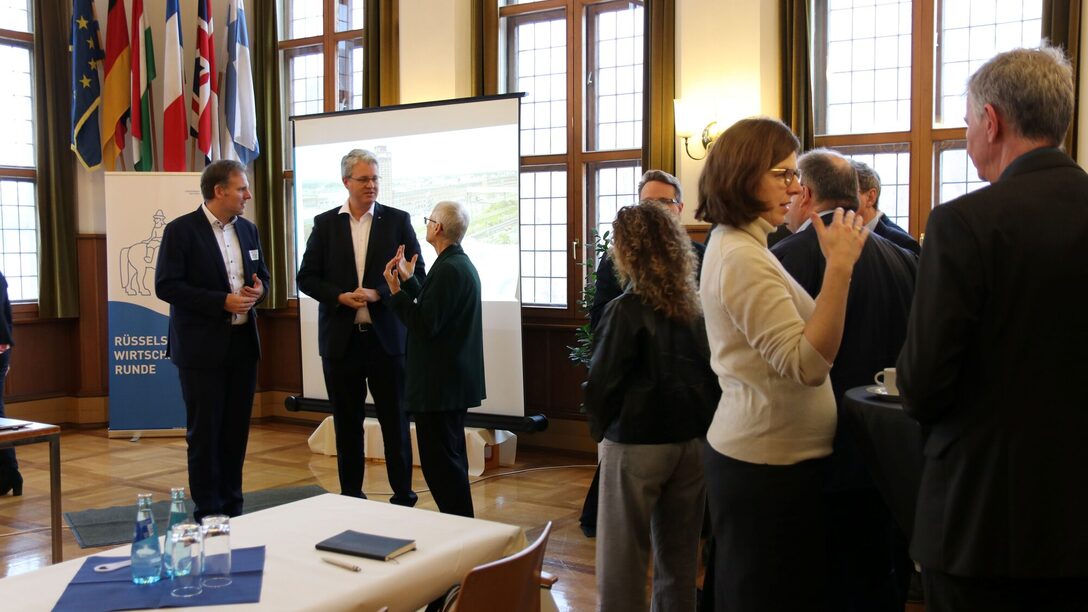 Blick in den Trausaal, in dem Teilnehmende der Wirtschaftsrunde in lockeren Grüppchen zusammenstehen und plaudern. Vor der Leinwand sind Maik Landwehr, Prof. Dr. Birgit Scheppath und OB Burghardt in ein Gespräch vertieft.