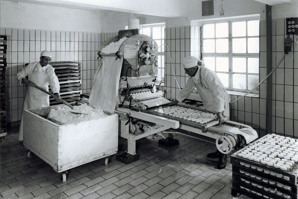 Schwarz-weiß Fotografie einer Formmaschine für Handkäse. Zwei weiß gekleidete Herren bedienen diese.